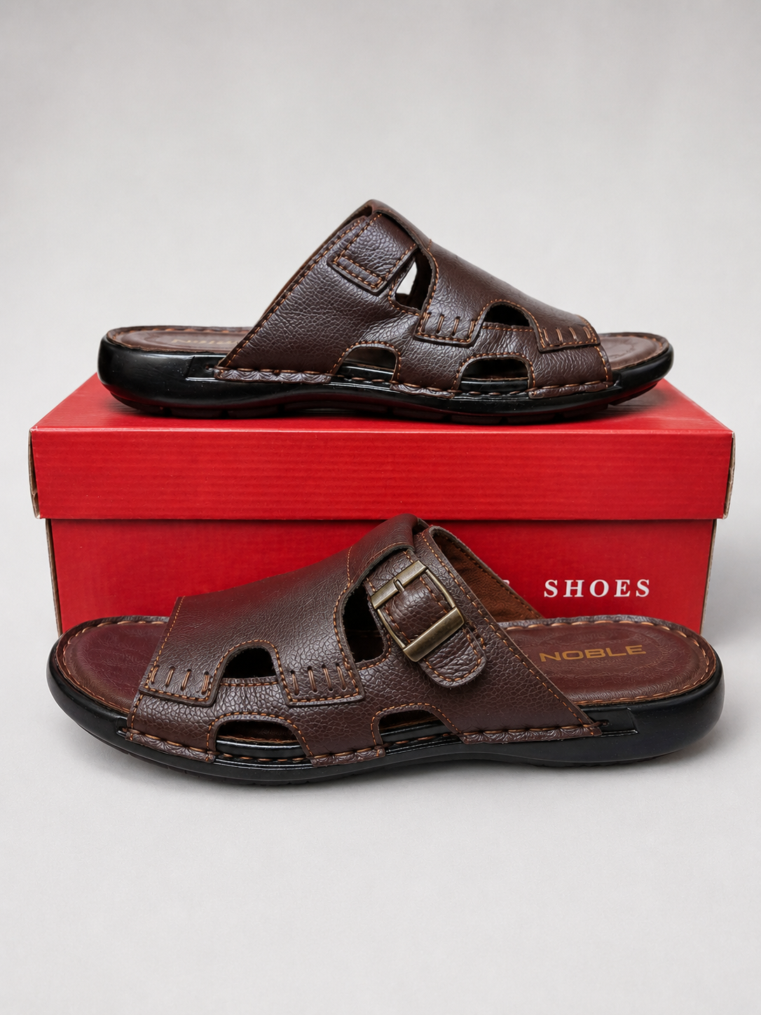 A pair of dark brown leather men's slide sandals with double straps, displayed on a red shoebox against a light gray background. The brand name 'NOBLE' is visible on the insole of the lower sandal.