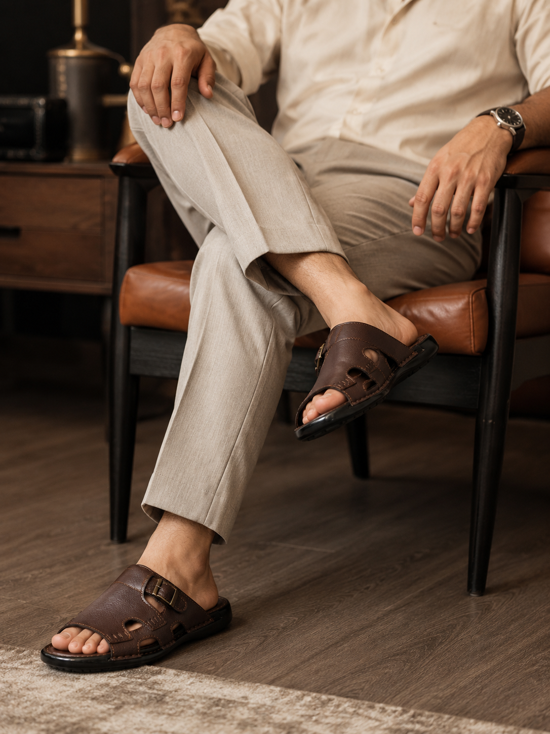 A man seated in a brown leather chair wearing dark brown leather slide sandals with adjustable straps and beige trousers in a warm indoor setting.